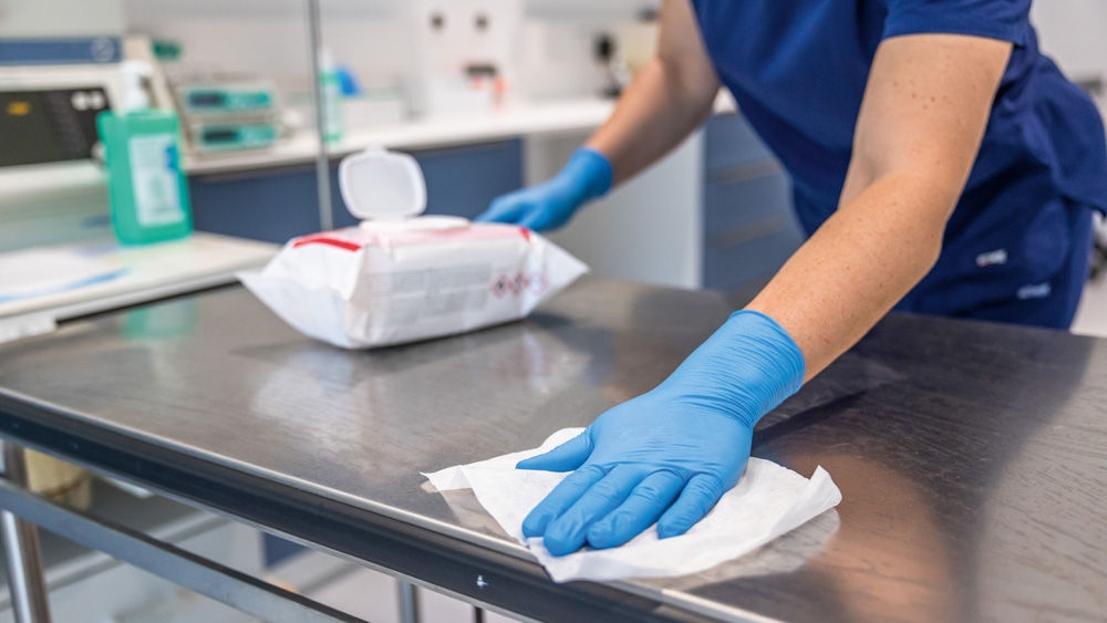 Veterinary assistant wearing blue nitrile gloves (Vasco Nitril Sky blue) cleaning a table with a disinfectant wipe (Meliseptol Wipes Sensitive) in a veterinary clinic.