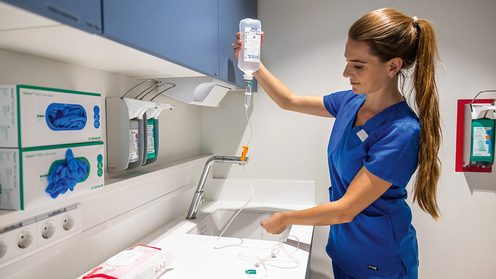 Veterinary professional in blue scrubs preparing an intravenous fluid infusion, holding an IV bag and tubing over a clinical work surface in a treatment area with medical supplies and equipment.