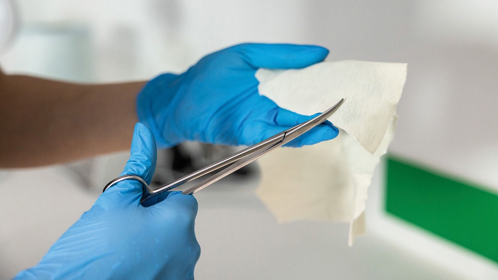 A veterinarian wearing Vasco Nitril gloves cuts a piece of white gauze or nonwoven material with AESCULAP surgical scissors.