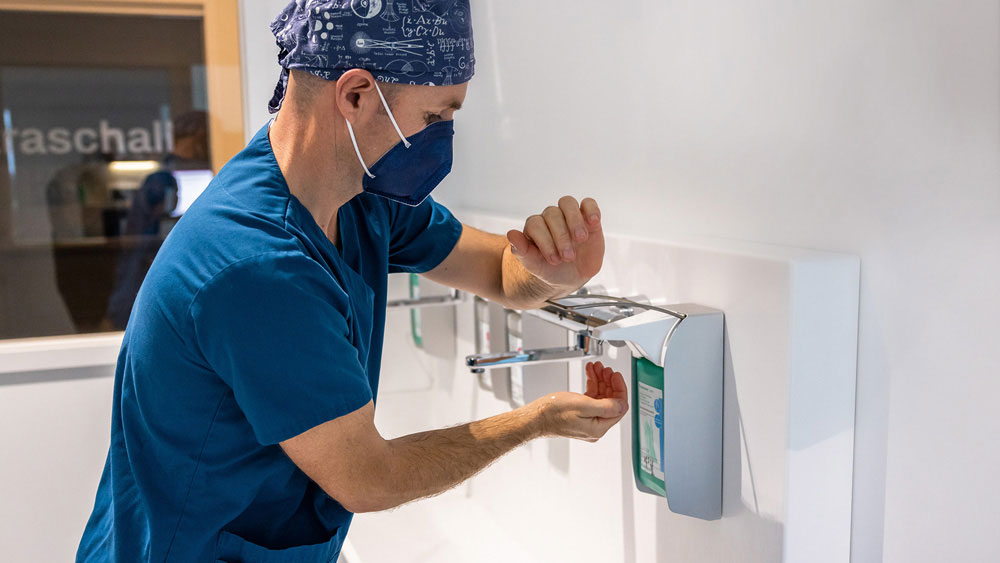 A veterinarian in surgical attire using a wall-mounted dispenser to apply hand disinfectant.