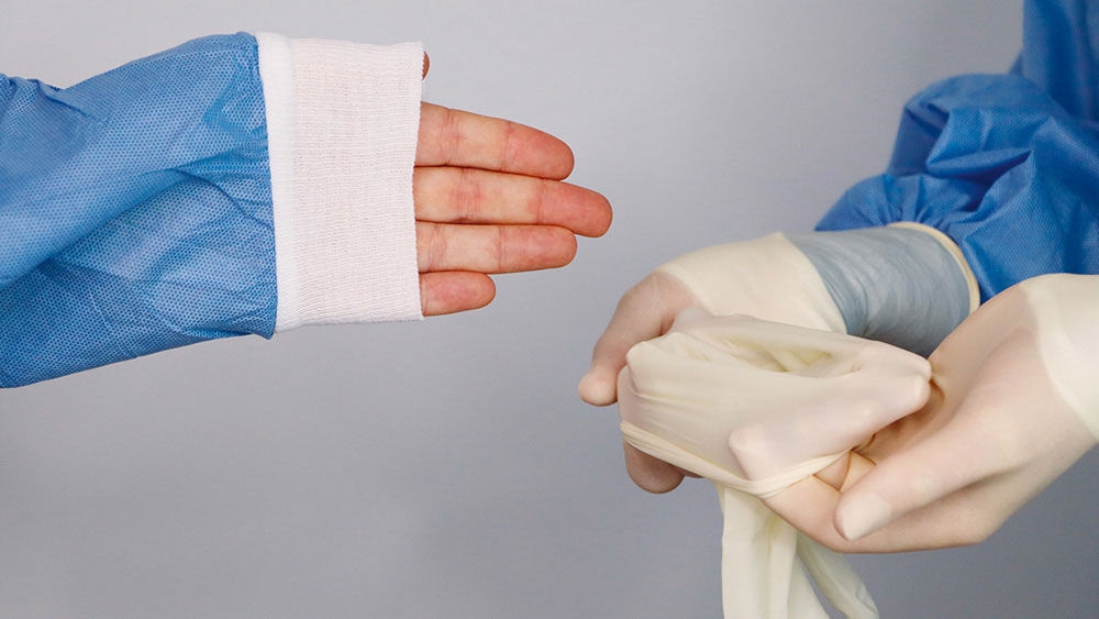 A gloved hand holding an open sterile glove while another person in a blue surgical gown extends their hand to put it on medical work surface.
