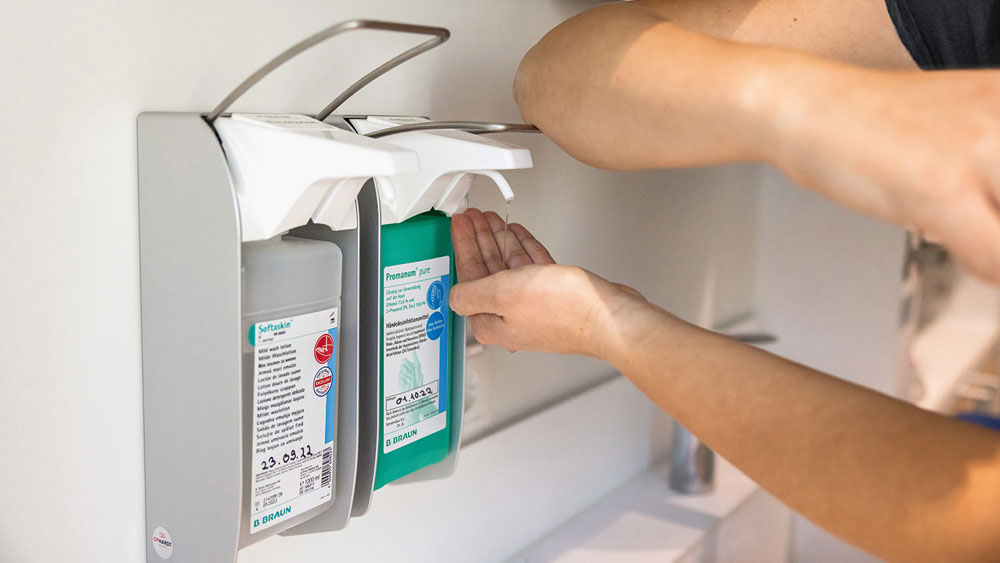 A veterinary nurse dispensing hand sanitizer from a wall mounted dispenser, holding their hands beneath the pump to apply the disinfectant.