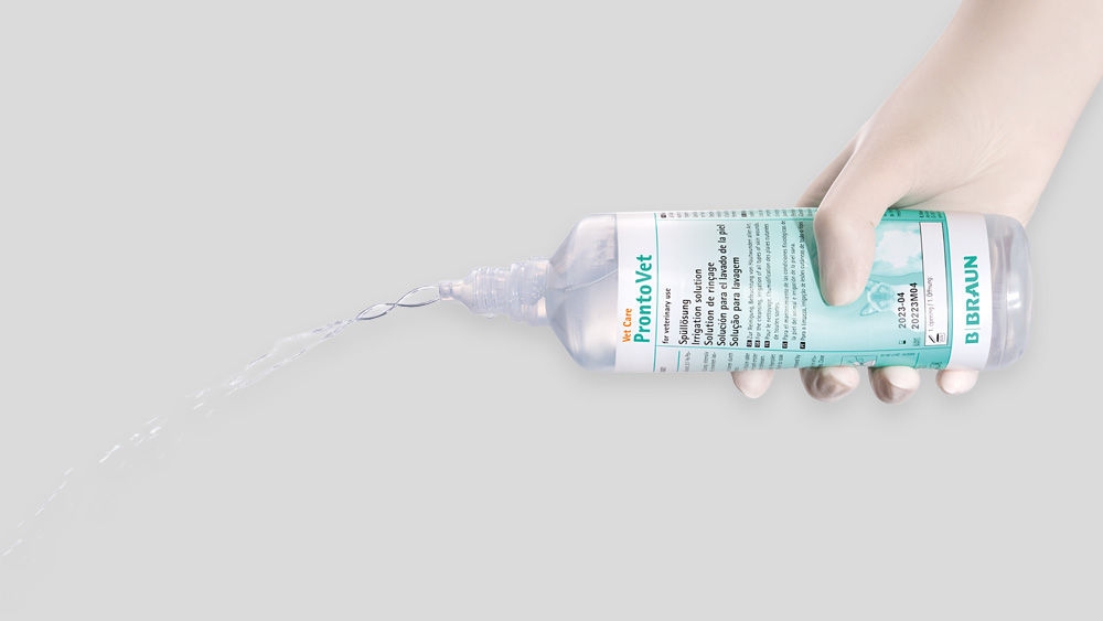 A hand holds a ProntoVet irrigation bottle from B. Braun at an angle, allowing a steady stream of liquid to pour out. The background is light grey.