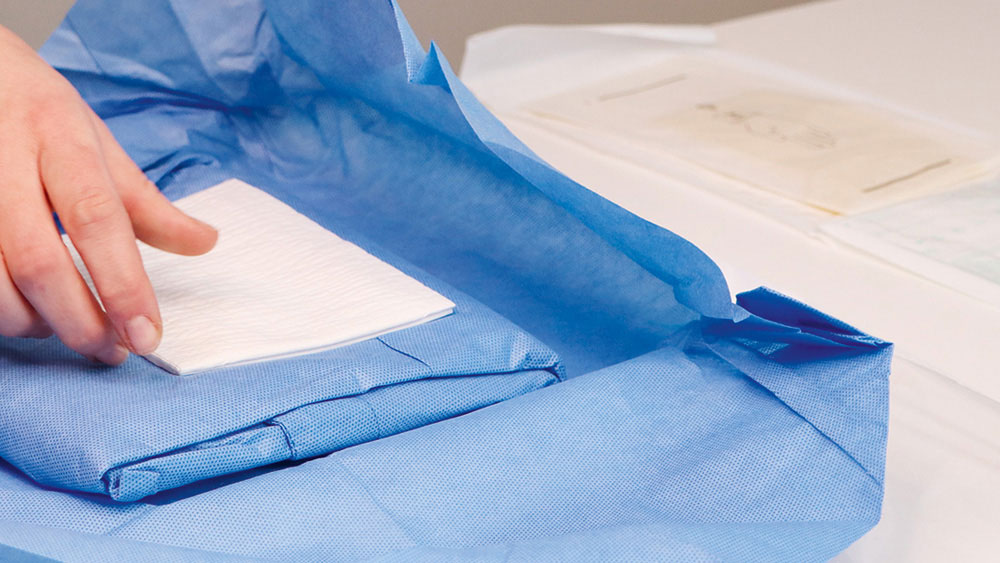 A hand placing a white cloth onto an opened blue sterile drape being prepared on a medical work surface.