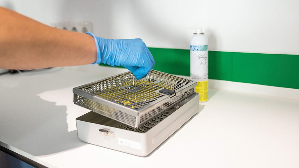 A veterinarian wearing blue disposable gloves lifts a metal sterilization tray filled with surgical instruments from a container on a clean countertop.