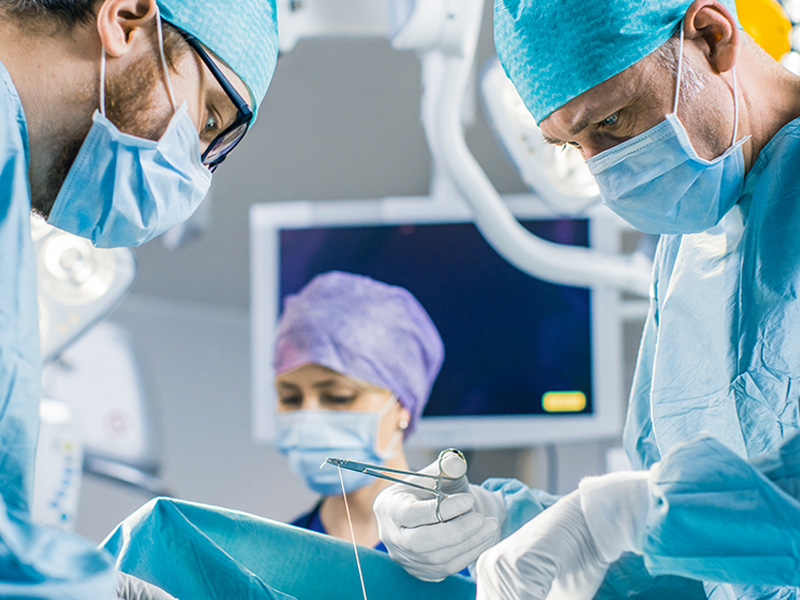 Medical staff in an operating room performing a surgical procedure, with gloved hands holding instruments and a monitor visible in the background.