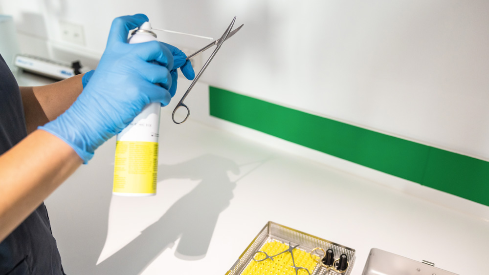 Gloved hands holding surgical scissors while applying disinfectant spray in an instrument reprocessing area.