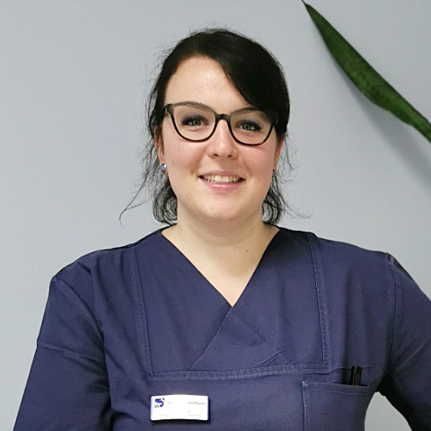 Veterinary assistant wearing dark medical scrubs standing indoors near a plant.