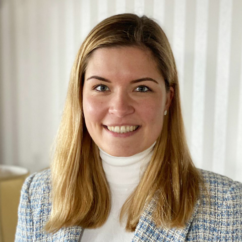 Person wearing a light-colored turtleneck and a checked blazer, standing indoors in front of vertical curtains.